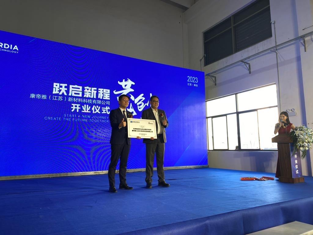 Two people holding a white board on a stage with a blue screen behind them, at the official opening of the new production facility of Concordia Textiles in Haian, China.
